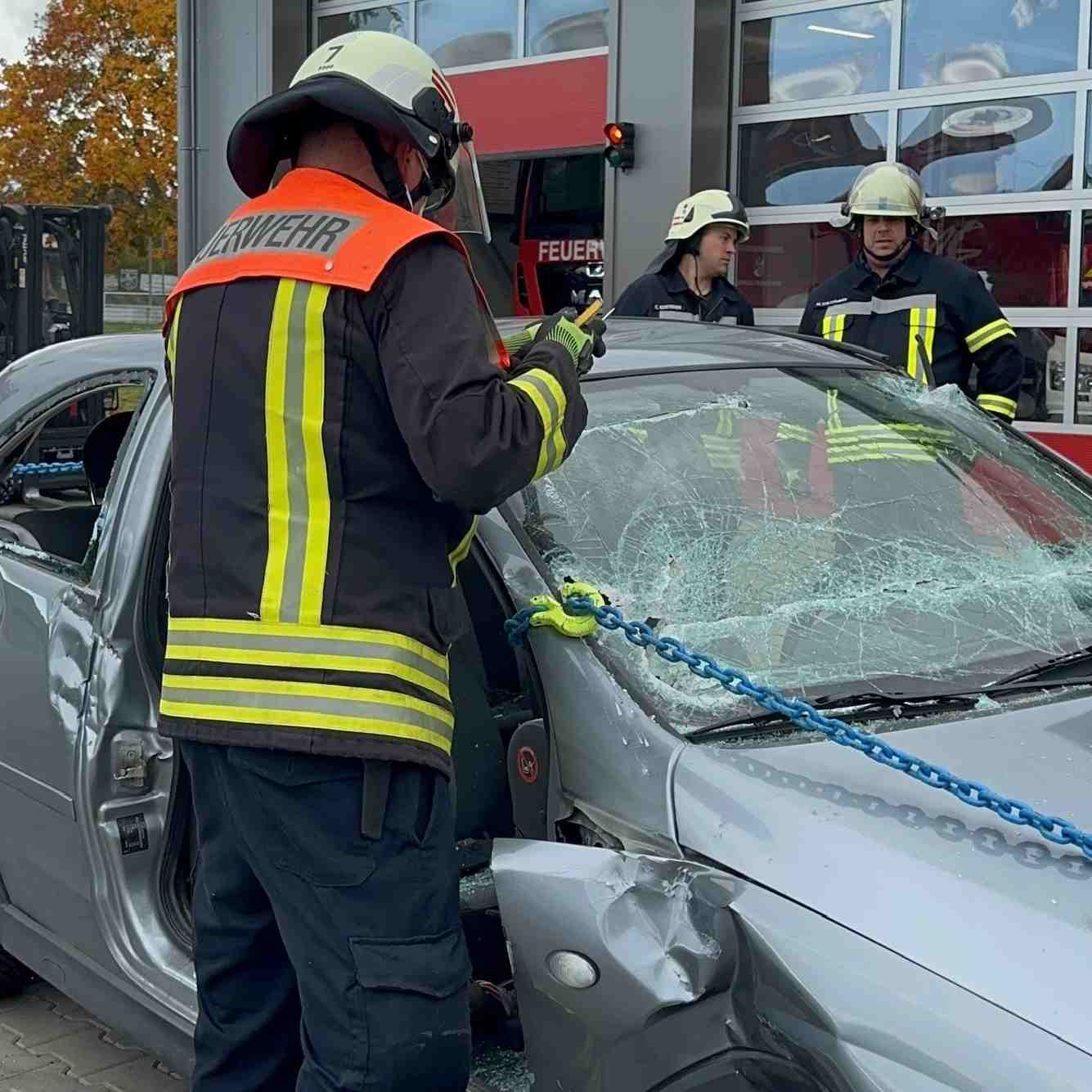 Drei Feuerwehrmänner in Schutzkleidung stehen bei einem verunfallten Auto mit zerbrochenem Fenster und eingedellter Karosserie. Kettenrettung nach der Oslomethode - Training Technical Resuce RH