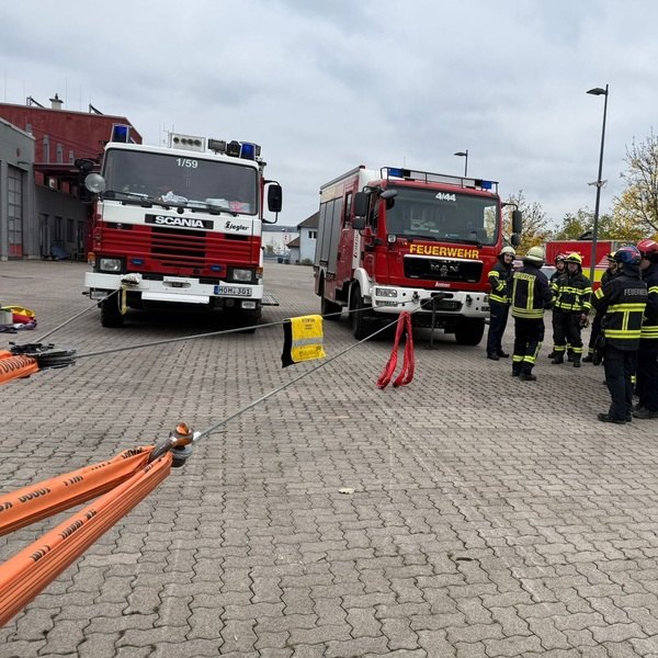 Zwei rote Feuerwehrfahrzeuge nebeneinander auf gepflastertem Hof, davor Feuerwehrleute in Uniformen und Helmen stehend mit orangefarbenen Schläuchen im Vordergrund. Maschinelle Zugeinrichtung Seminar