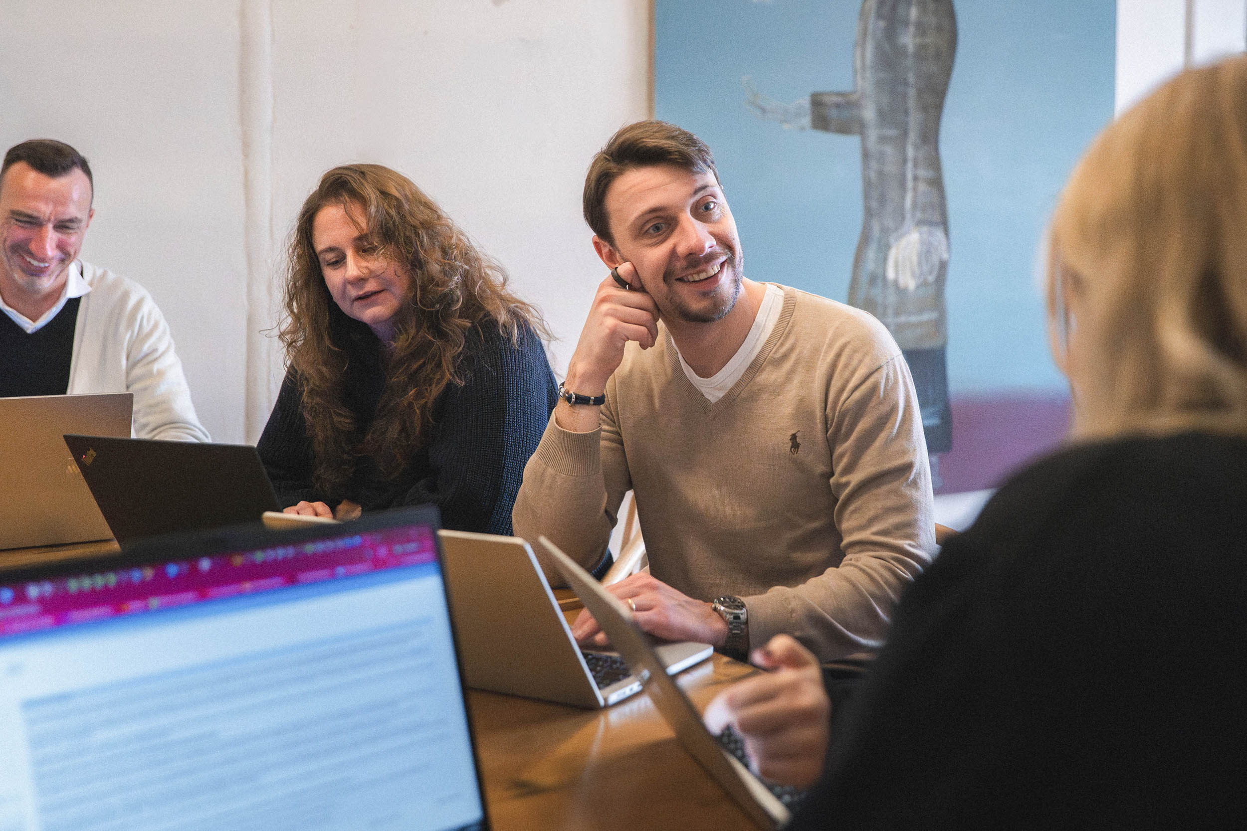 Eine männliche Person unterhält sich mit seinen Kolleg:innen die am Laptop arbeiten über modernes Arbeitsrecht im digitalen Zeitalter