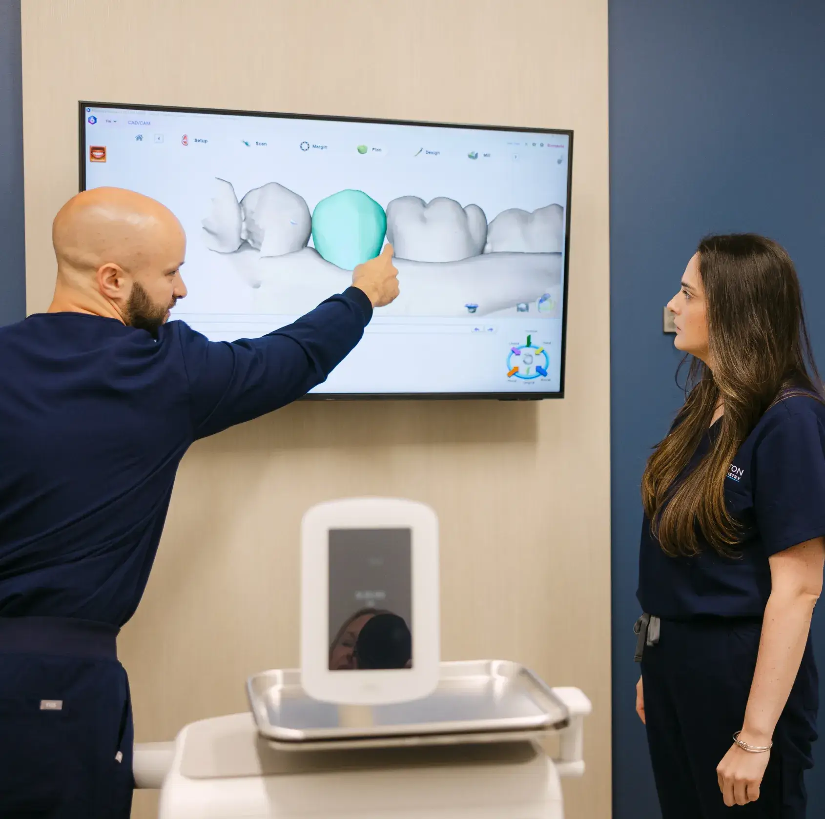 A man in scrubs points at a dental scan on a screen while a woman listens.