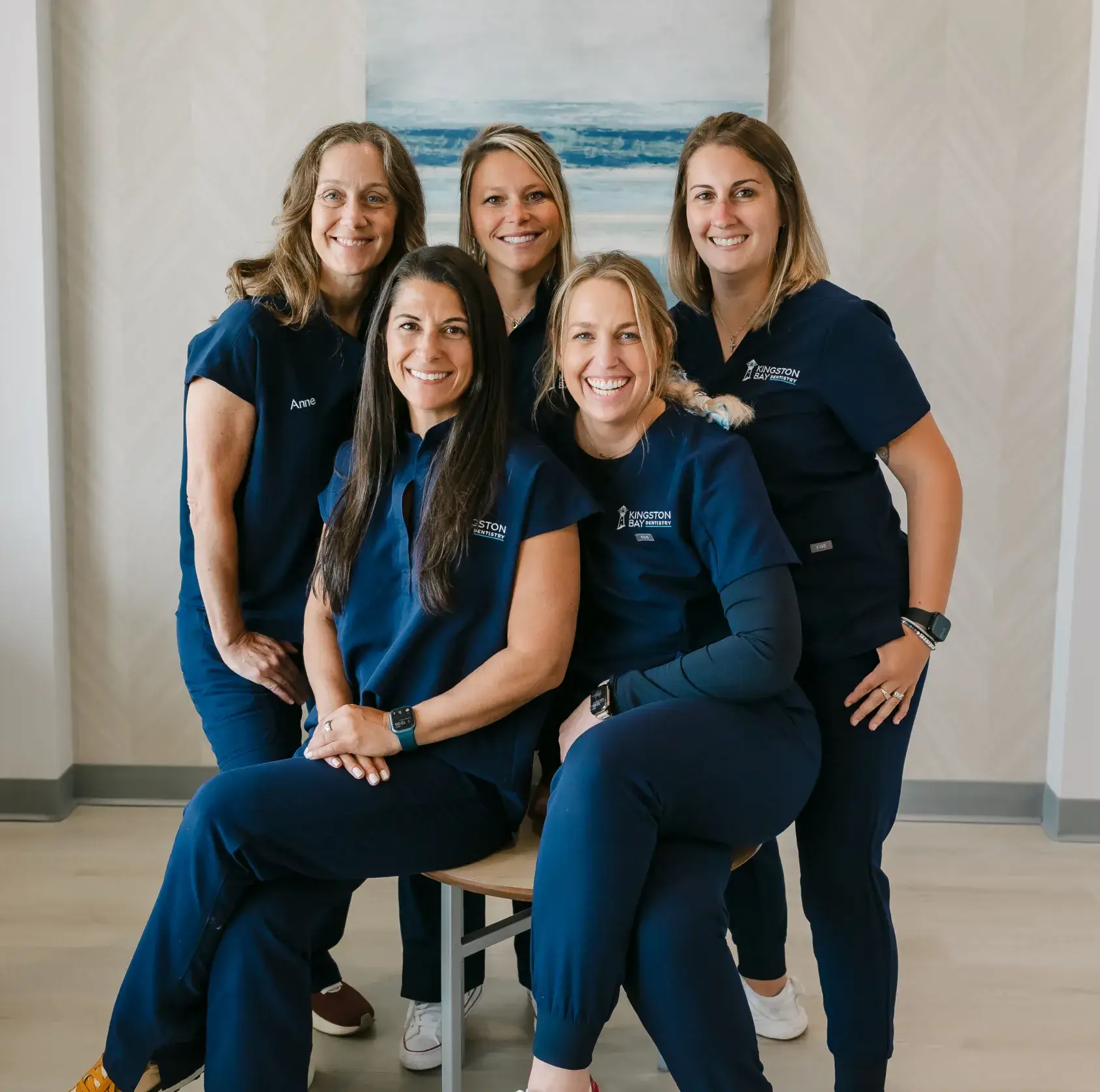 Five women wearing blue medical uniforms smile and pose together in a well-lit room.