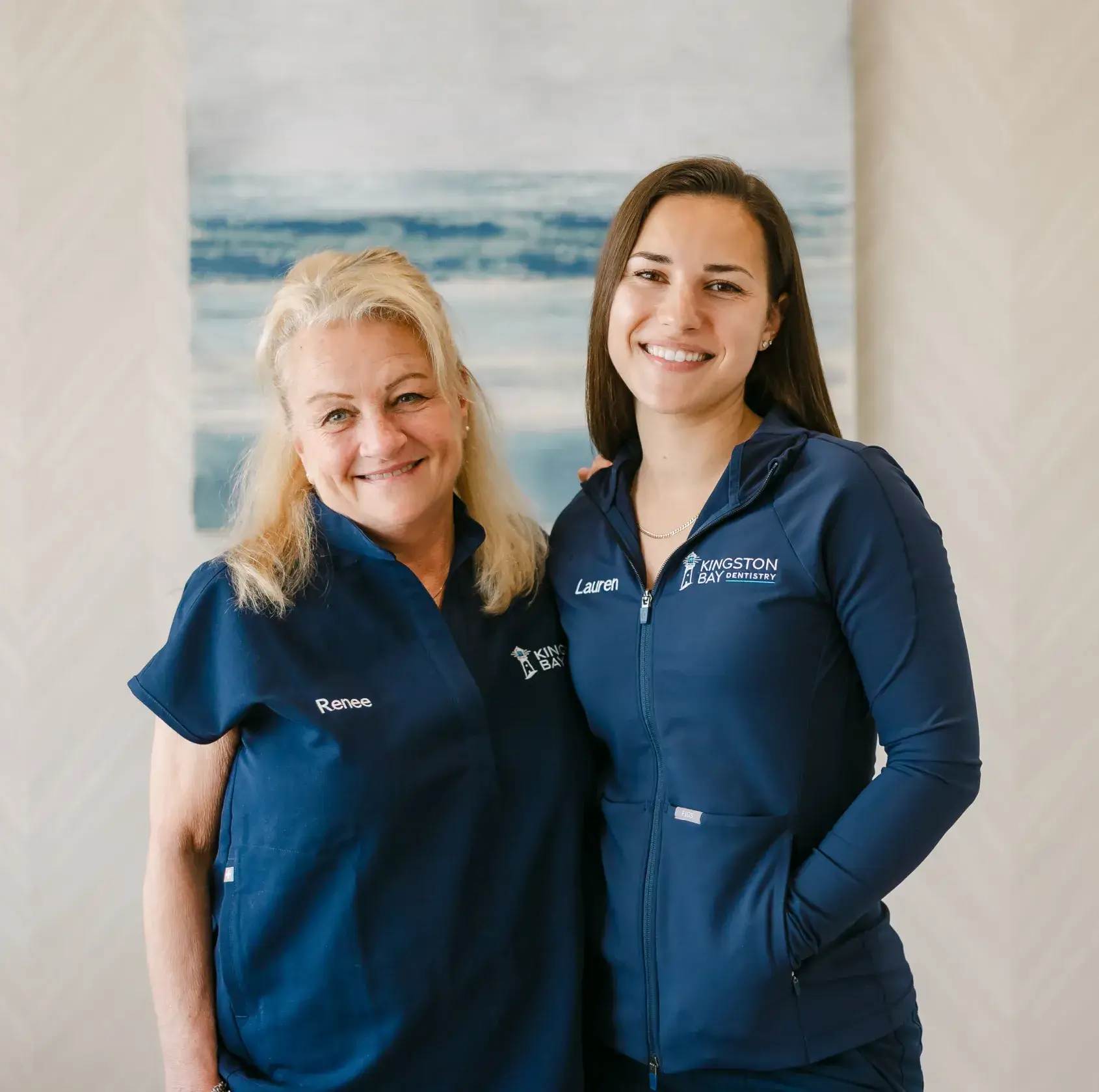 Two women wearing matching navy uniforms, smiling and standing next to each other indoors.