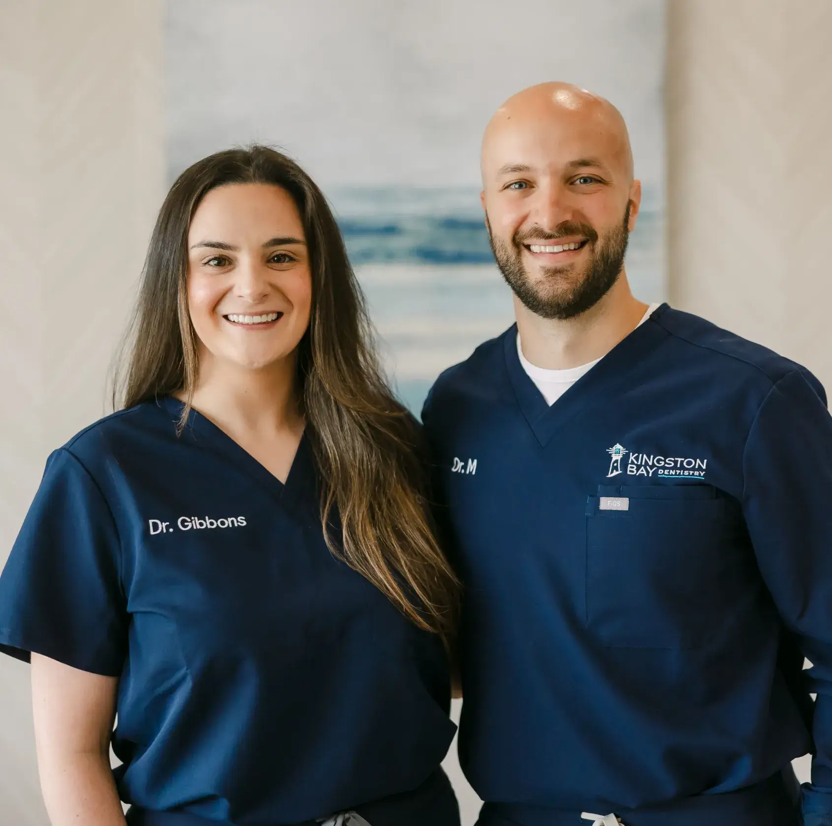 Two dentists in navy scrubs smiling, standing side by side in an office setting.