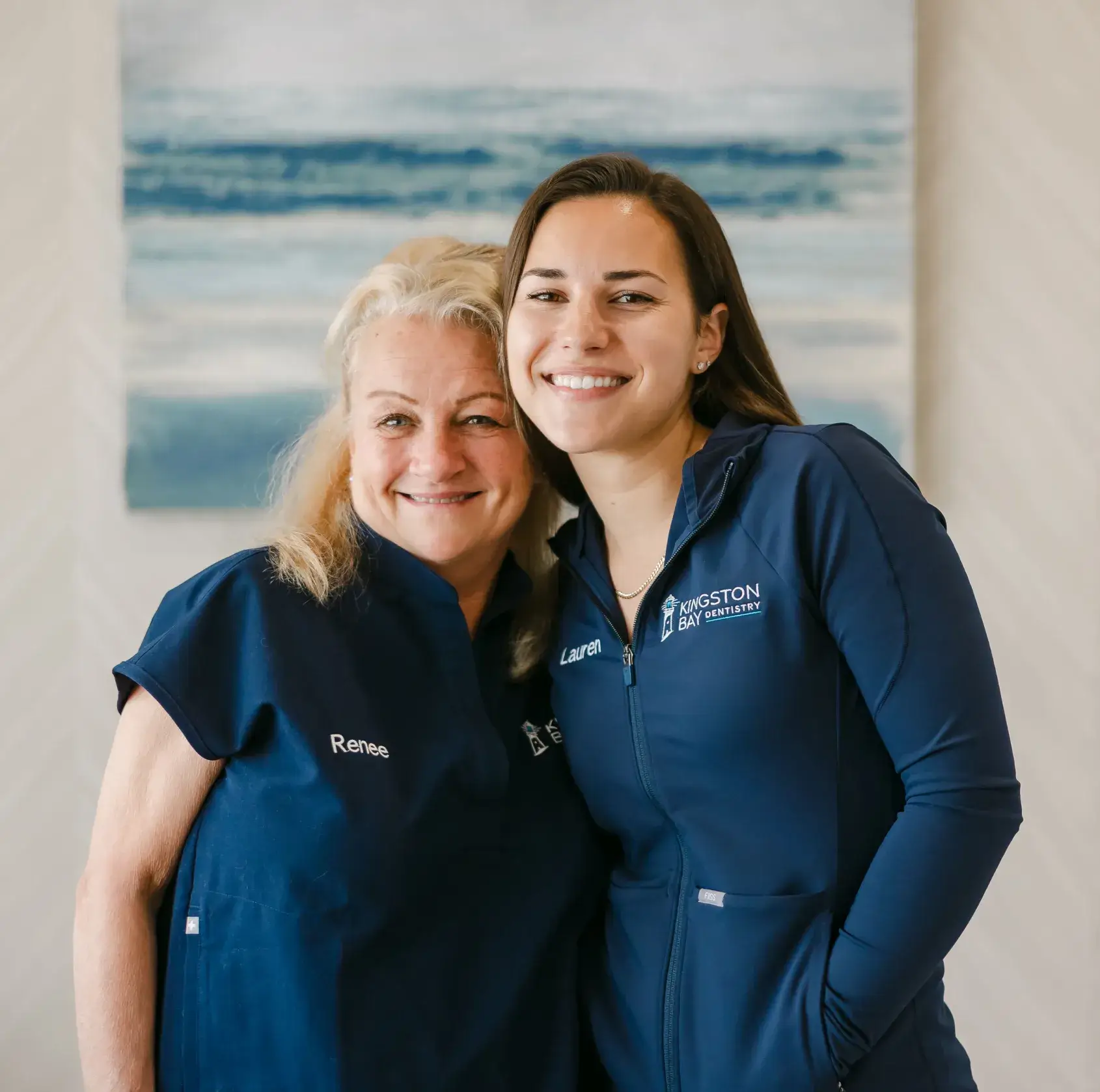 Two smiling women wearing matching blue uniforms stand closely together indoors.