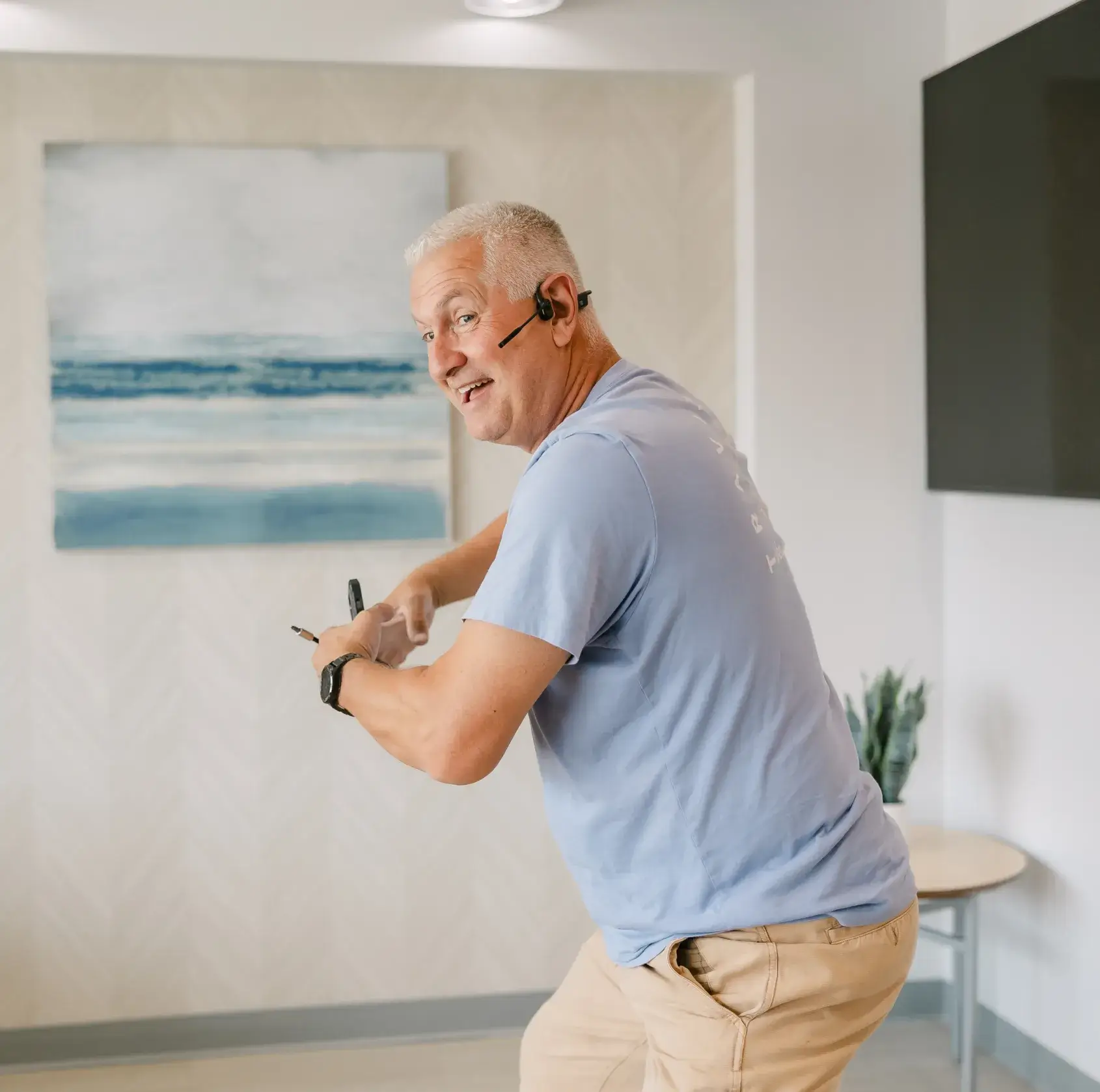 A person wearing a headset holds two pens, standing in a room with a painting.