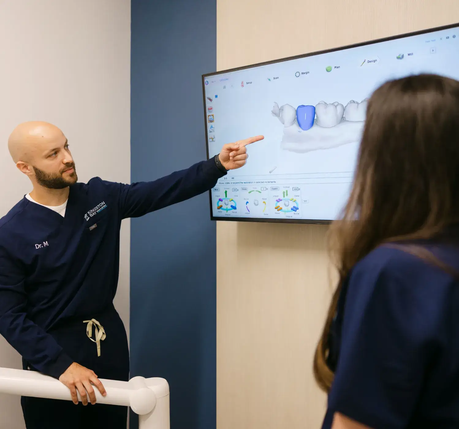 A dentist in blue scrubs points at a dental X-ray on a screen for a patient.