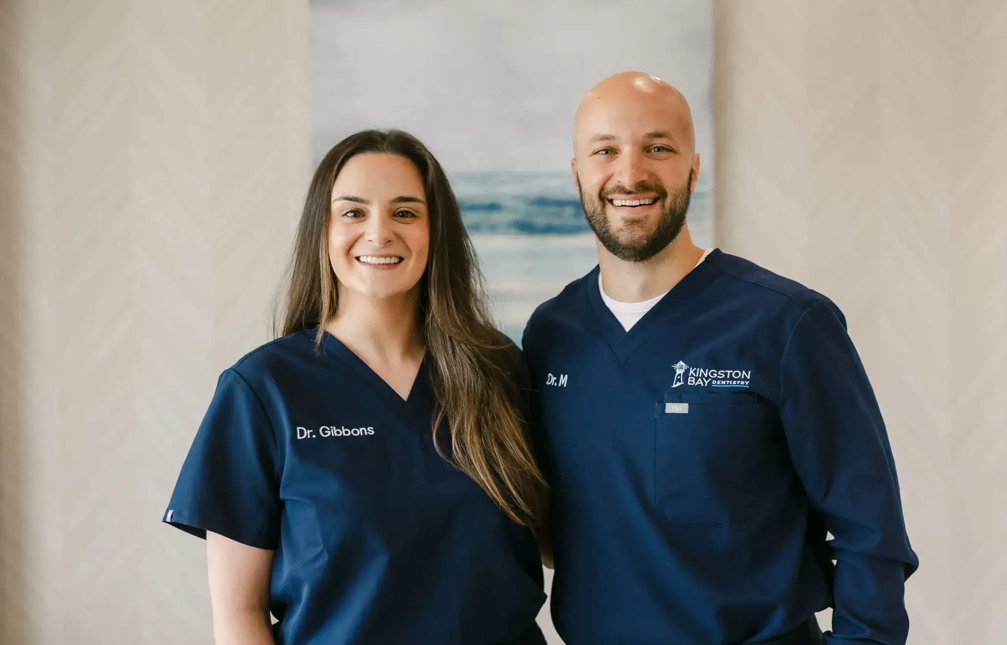 Two dental professionals smiling, wearing navy scrubs with name tags, standing indoors.