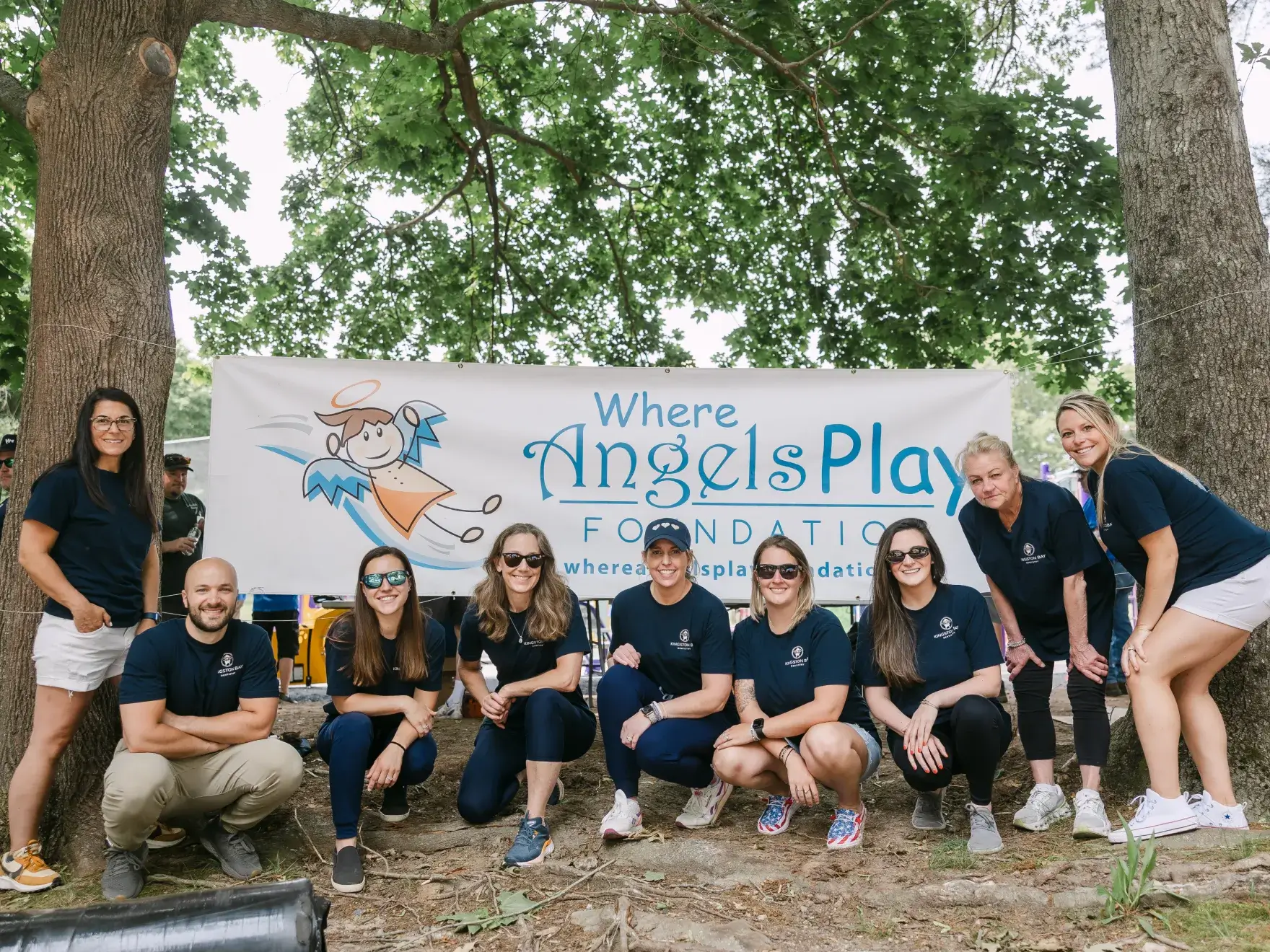 A group of people in matching shirts kneel and stand in front of the "Where Angels Play" banner.