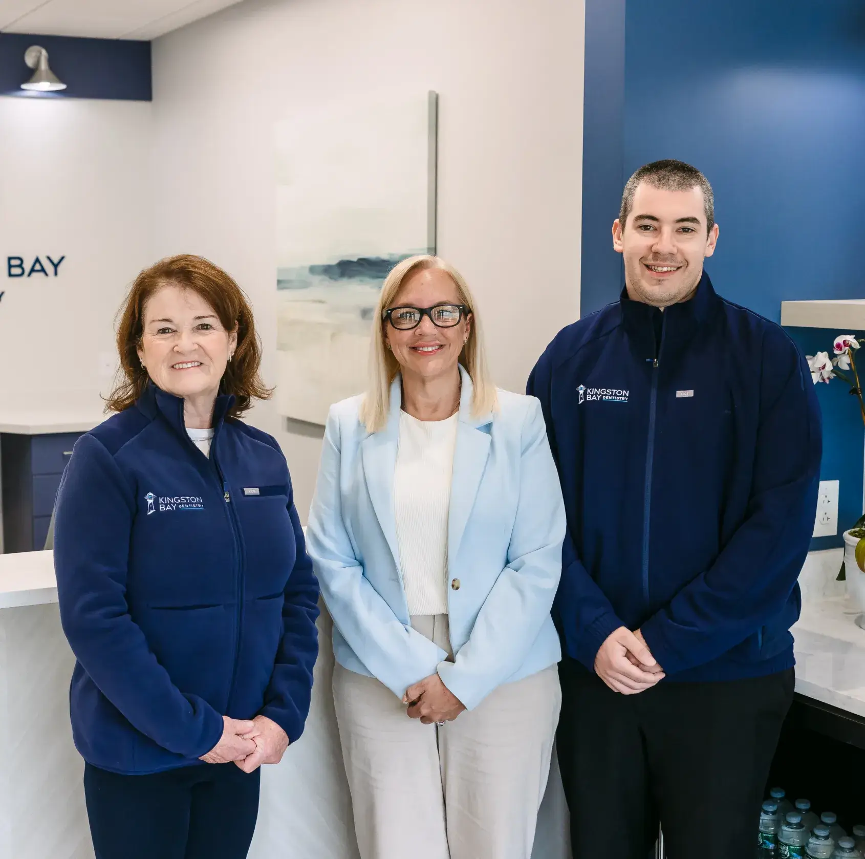 Three people are standing and smiling in an office, wearing business casual clothing.