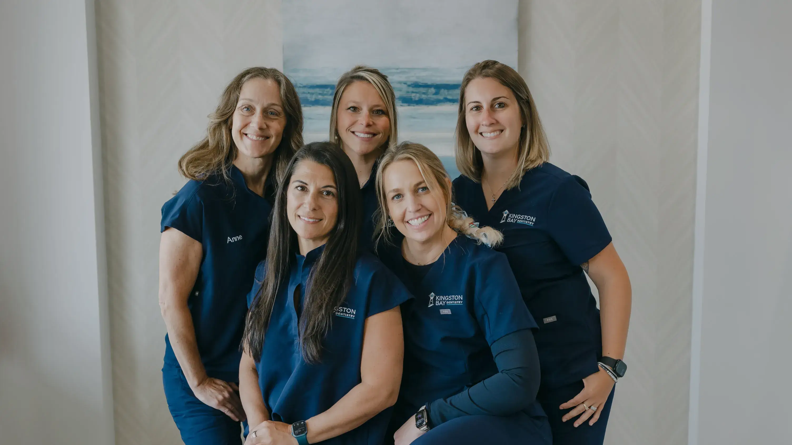 Five smiling women in matching blue uniforms pose together in an indoor setting.