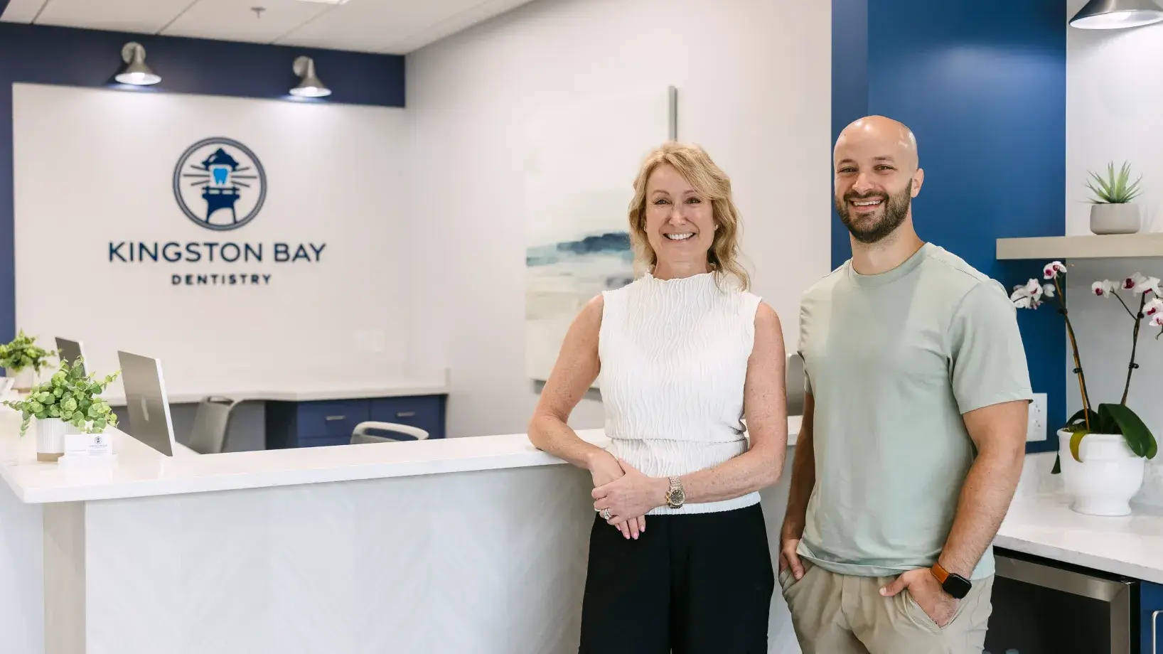 Two smiling people stand at a dental office reception desk, with the clinic's name visible on the wall.