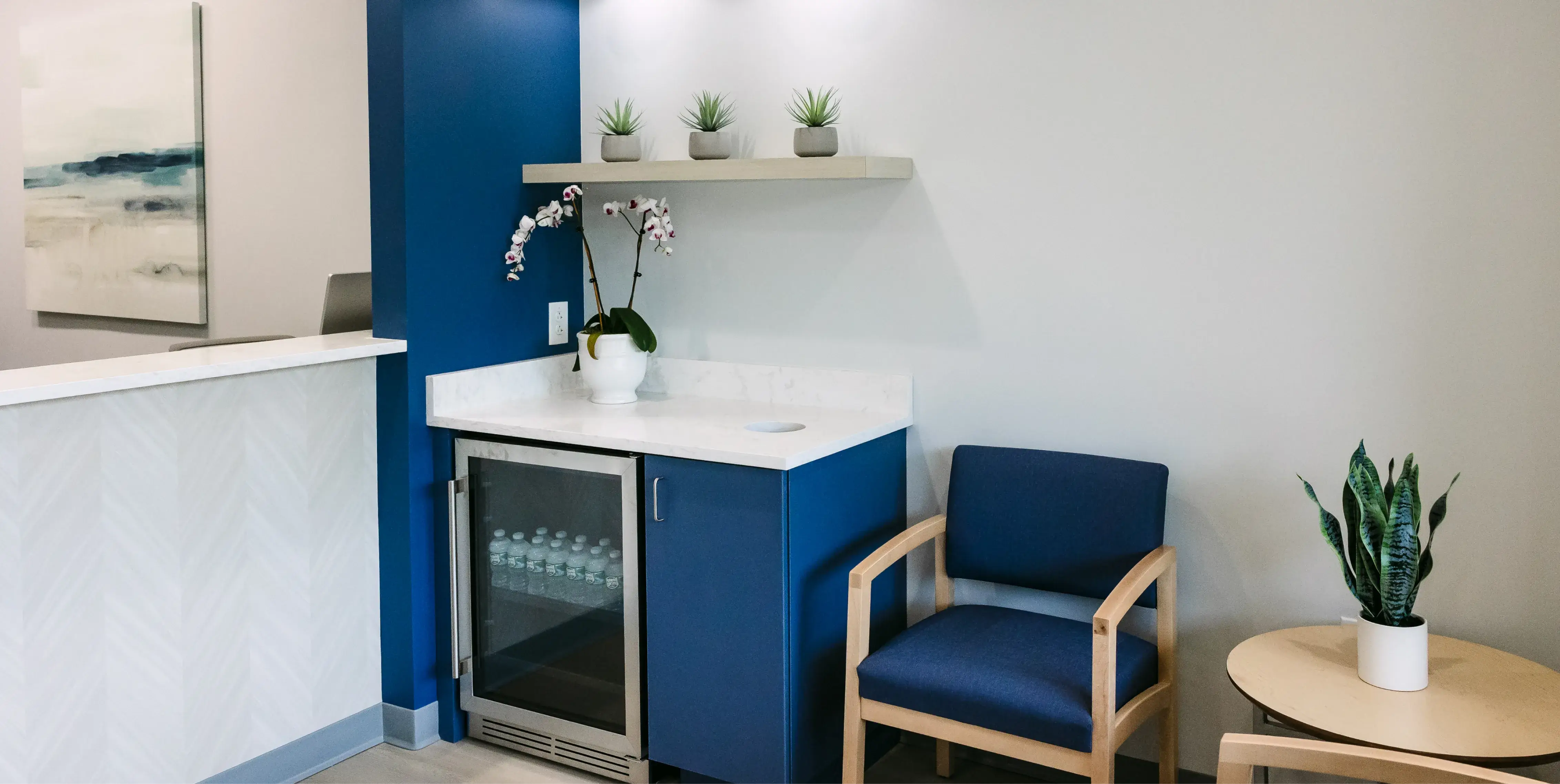 A waiting room with a blue chair, a small table, and a mini fridge with bottled water.