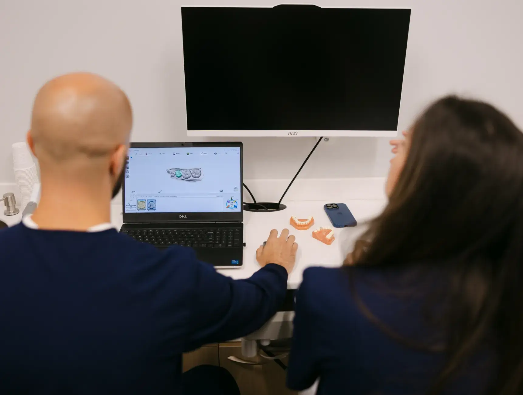 Two people sitting at a desk, looking at a laptop with dental images on the screen.