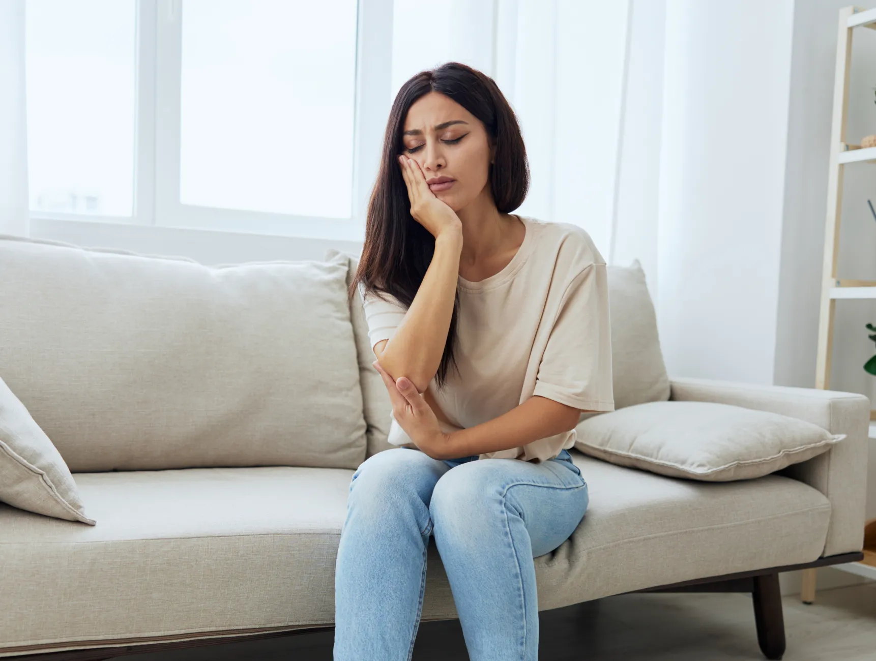 A woman sits on a couch, holding her cheek with a pained expression.