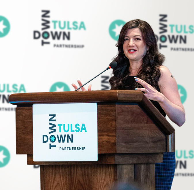 Photo of District 4 Tulsa City Councilor Laura Bellis gesturing as she speaks into a microphone at a lecturn with a Downtown Tulsa step-and-repeat banner behind her