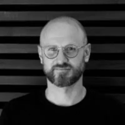 Portrait of a bald man with a beard and round glasses wearing a black shirt against a dark horizontal striped background.
