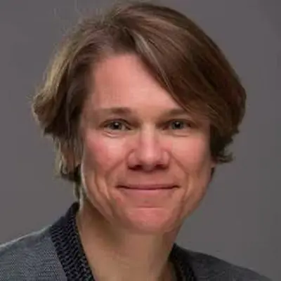 Portrait of a middle-aged person with short brown hair smiling gently against a gray background.