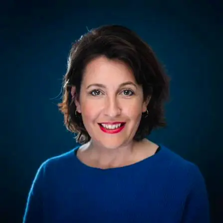 Smiling woman with short dark hair wearing a blue sweater and red lipstick against a dark blue background.