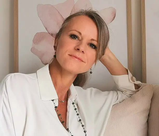 Smiling woman with light brown hair, wearing a white shirt, leaning on a beige pillow with a floral artwork background.