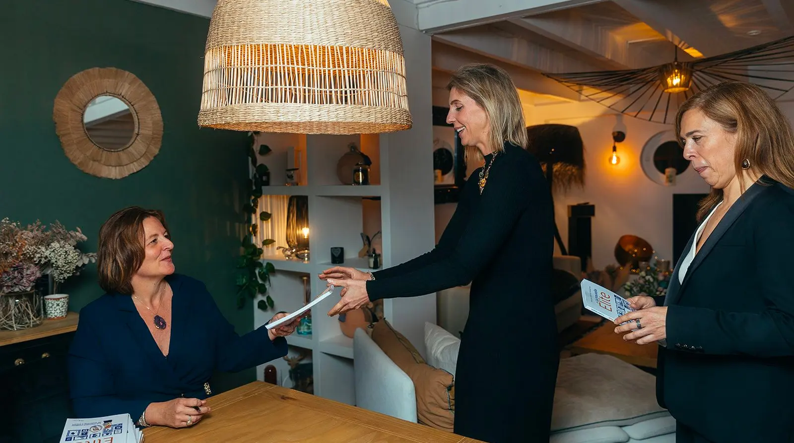 Three women indoors exchanging documents near a wooden table with decorative plants and warm lighting.