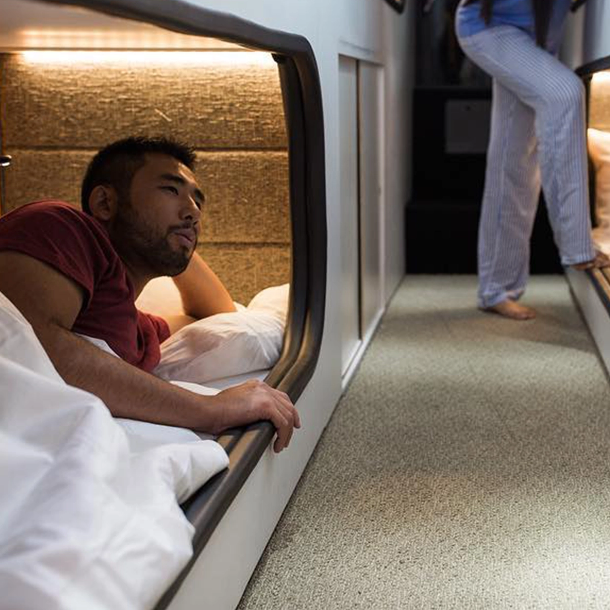 Passenger resting in a lower Cabin bunk as another walks the carpeted aisle