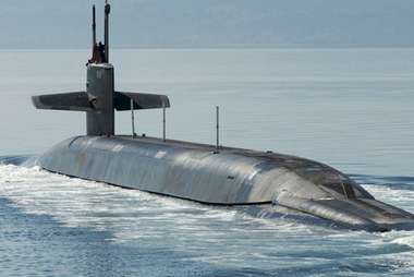 Military submarine partially surfaced and moving through calm ocean waters.