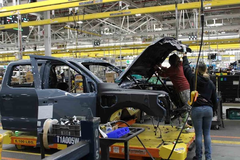 Two workers assembling a partially built black vehicle on a yellow platform inside a spacious automotive factory.