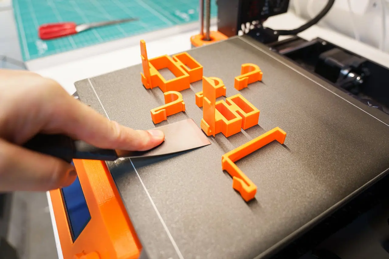 Hand using a metal scraper to remove bright orange 3D-printed parts from a textured PEI print bed, with a cutting mat and tools visible in the background.