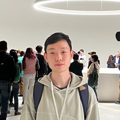 Young man wearing a beige hoodie and backpack standing indoors with a crowd behind him under circular light.
