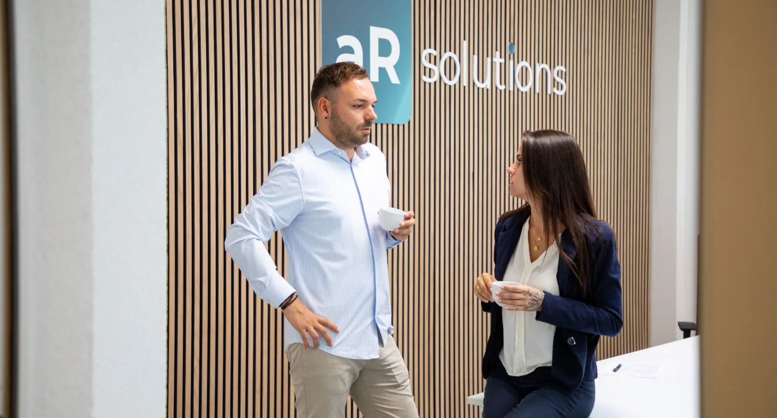 Zwei Personen stehen vor einer modernen Holzvertäfelung mit dem Logo „aR solutions“ und unterhalten sich bei einer Tasse Kaffee; professionelle Büroumgebung mit freundlicher Gesprächsatmosphäre.