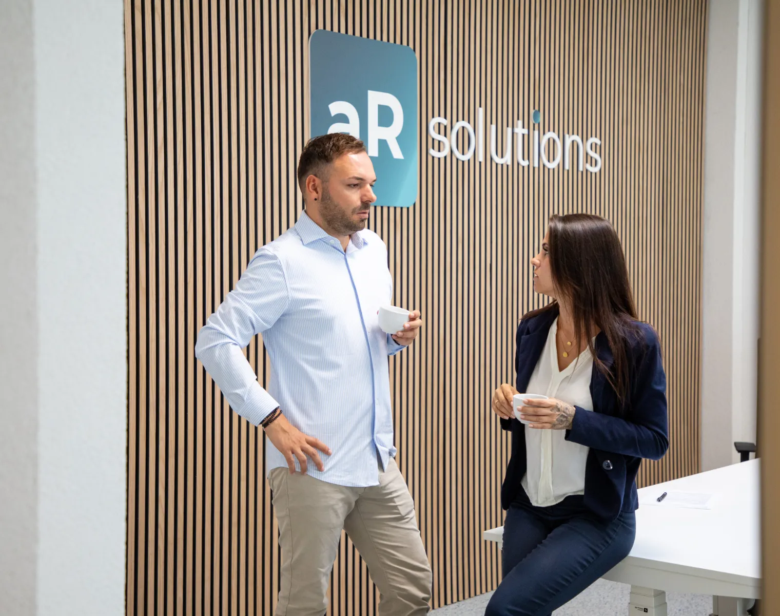 Zwei Personen stehen vor einer modernen Holzvertäfelung mit dem Logo „aR solutions“ und unterhalten sich bei einer Tasse Kaffee; professionelle Büroumgebung mit freundlicher Gesprächsatmosphäre.