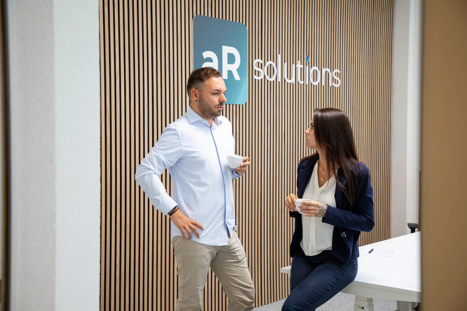 Zwei Personen stehen vor einer modernen Holzvertäfelung mit dem Logo „aR solutions“ und unterhalten sich bei einer Tasse Kaffee; professionelle Büroumgebung mit freundlicher Gesprächsatmosphäre.