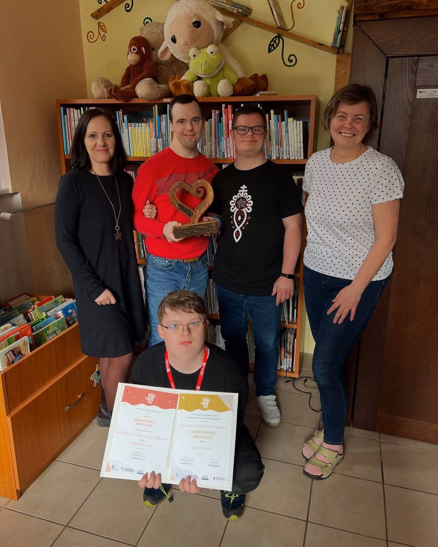 Skupina piatich ľudí v knižnici, dvaja držia certifikáty, jeden drevené srdce.