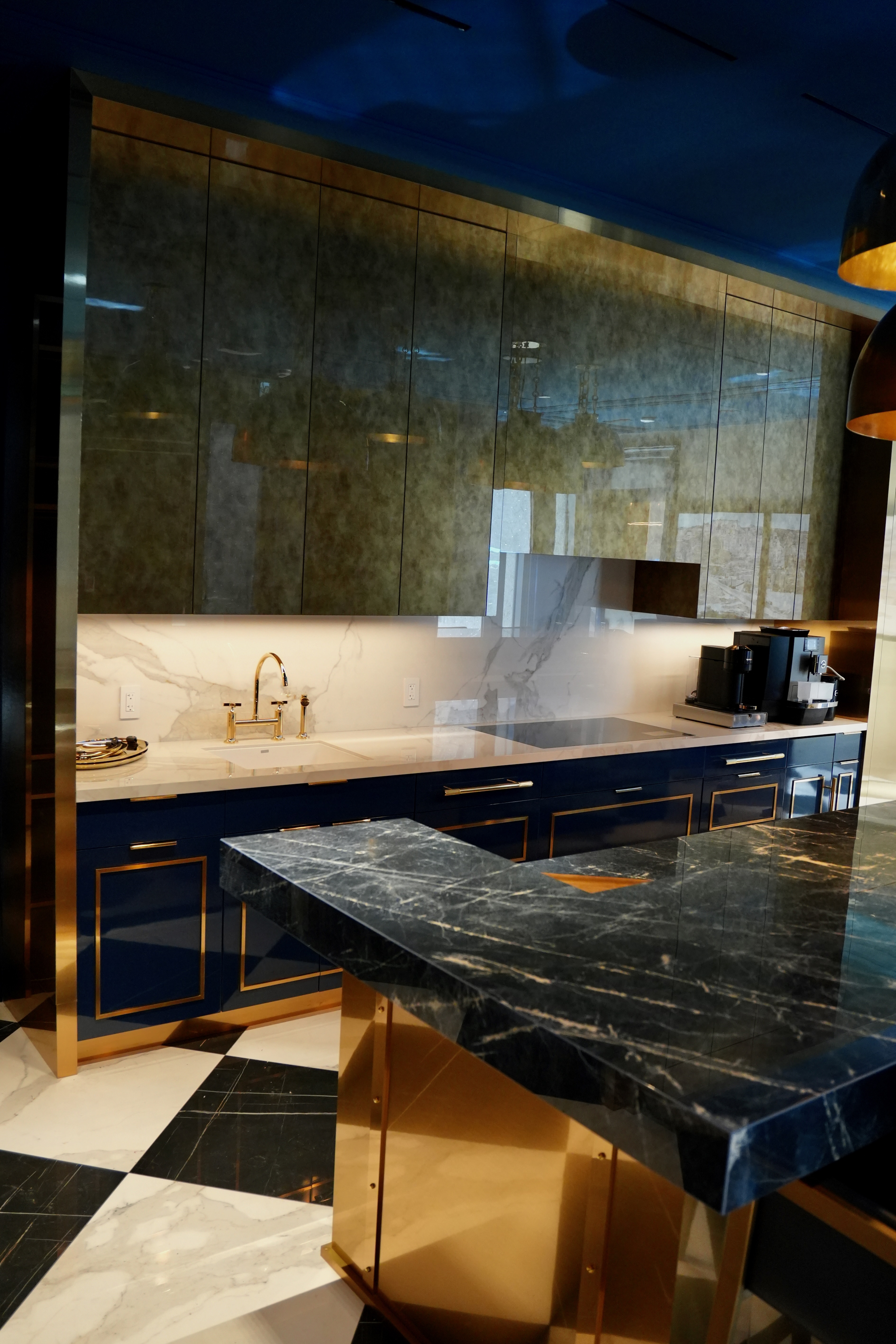 Modern kitchen with navy blue and gold-trimmed cabinets, white marble backsplash and countertop, black marble island with gold base, and coffee machine.