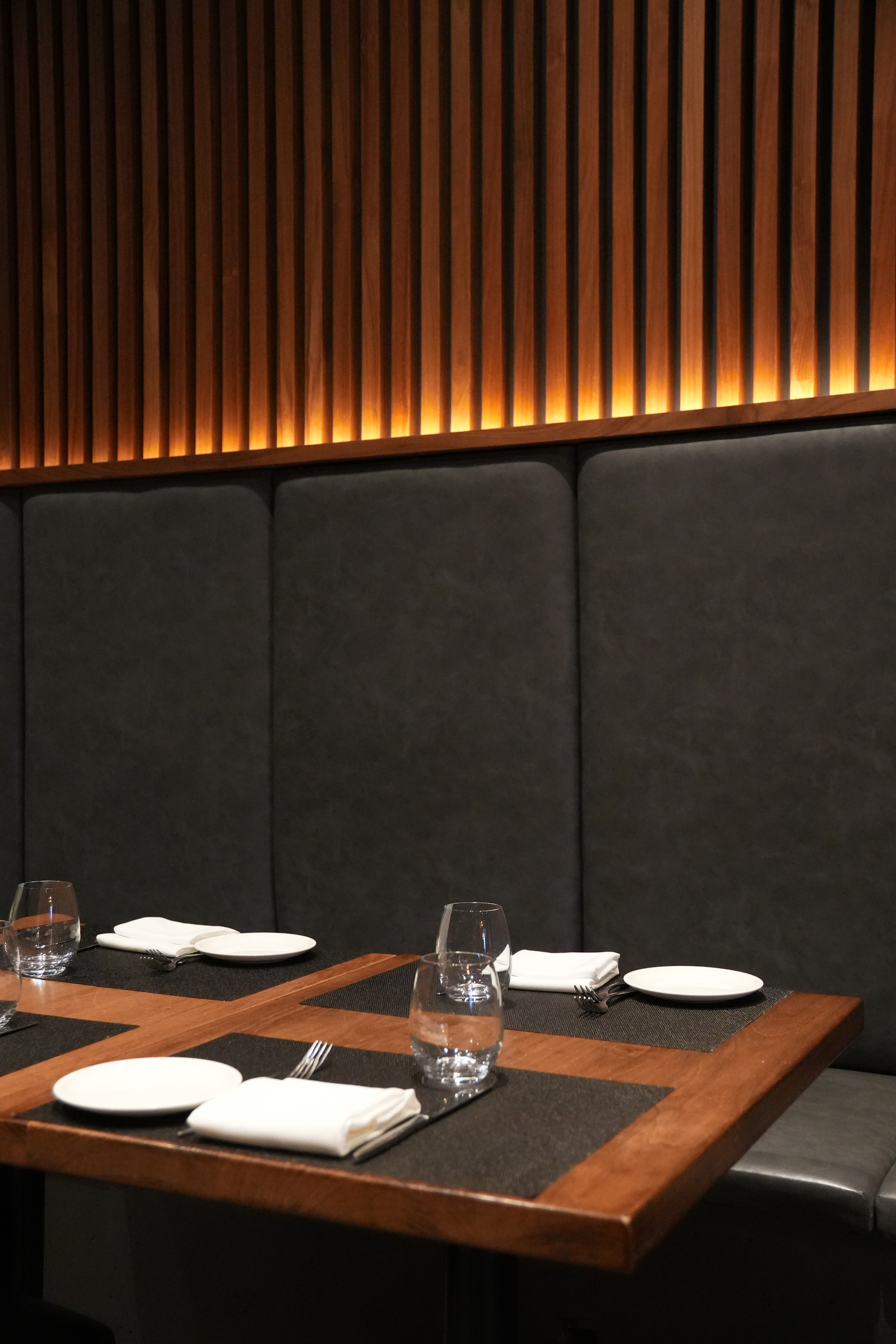 Modern restaurant booth seating with wood-paneled wall, set tables with black placemats, white plates, napkins, forks, and glasses.