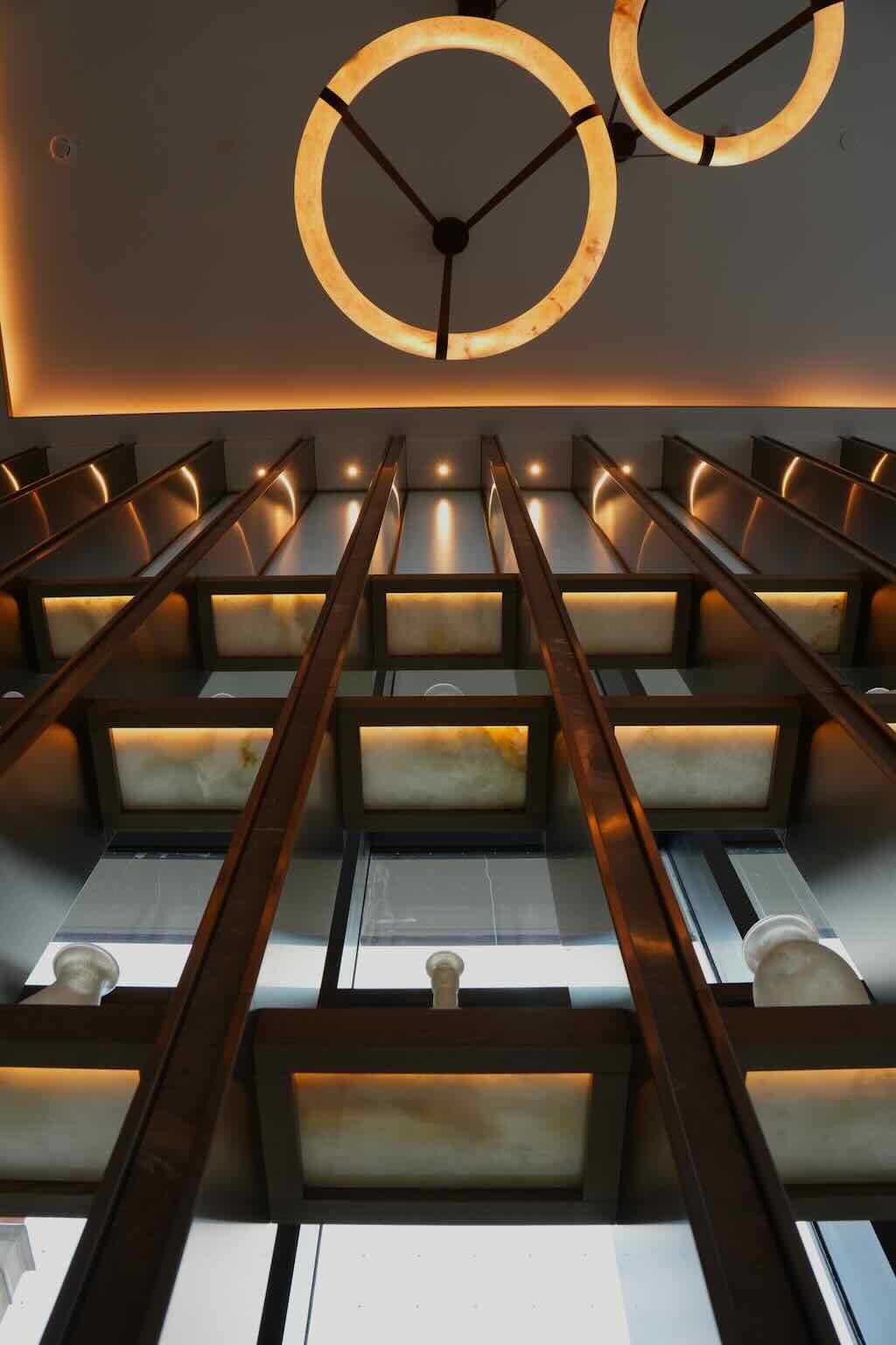 Modern ceiling light fixture with two illuminated circular rings and vertical wooden beams with backlit panels on the wall.