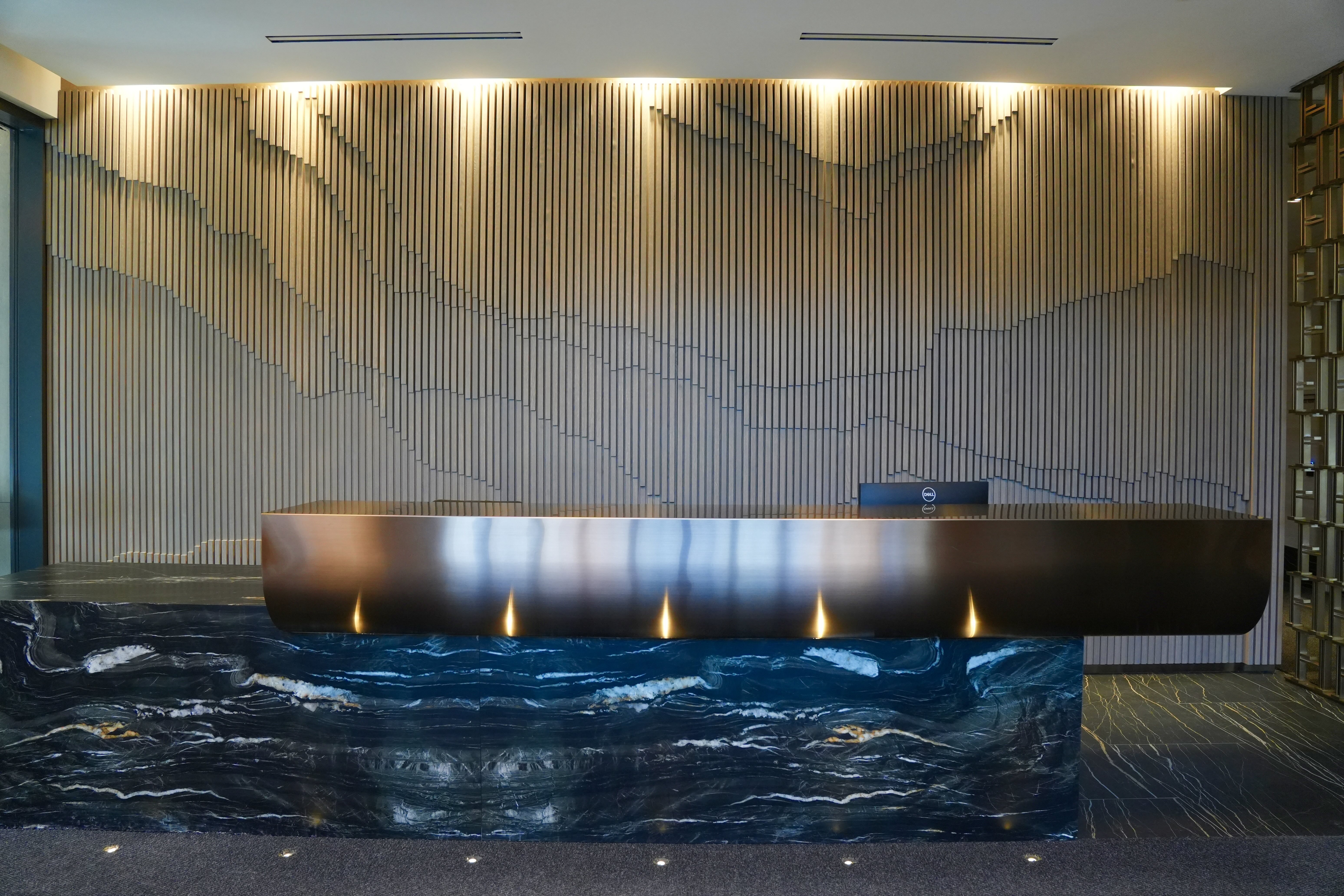 Modern reception desk with a sleek black marble base and a curved metallic front panel in front of a wooden slat wall with recessed lighting.