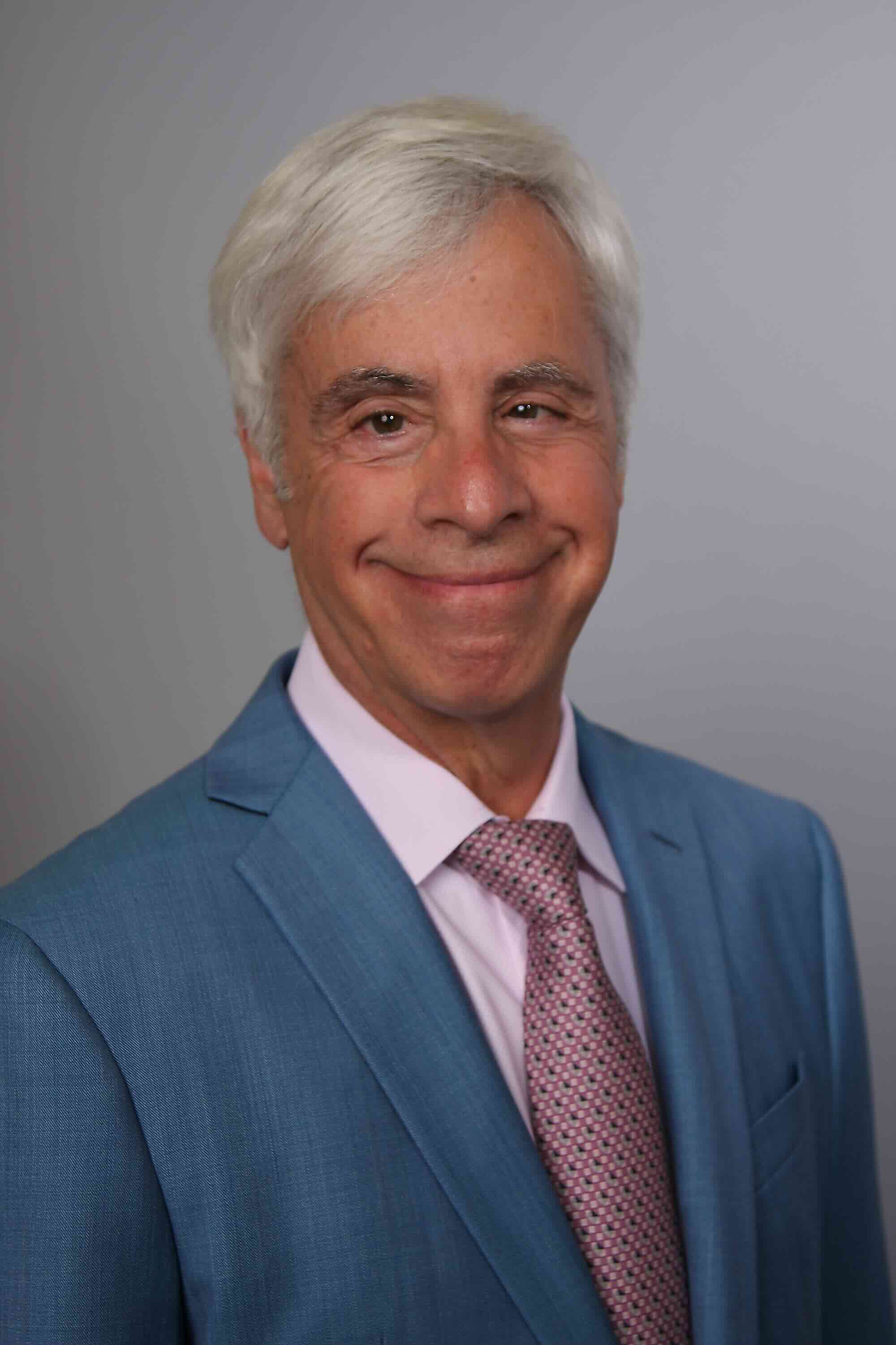 Audiologist with short gray hair wearing a dark suit, white shirt, and patterned tie against a gray background.
