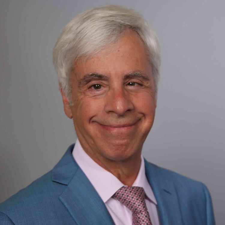Audiologist wearing a dark suit, white shirt, and patterned tie against a dark background.