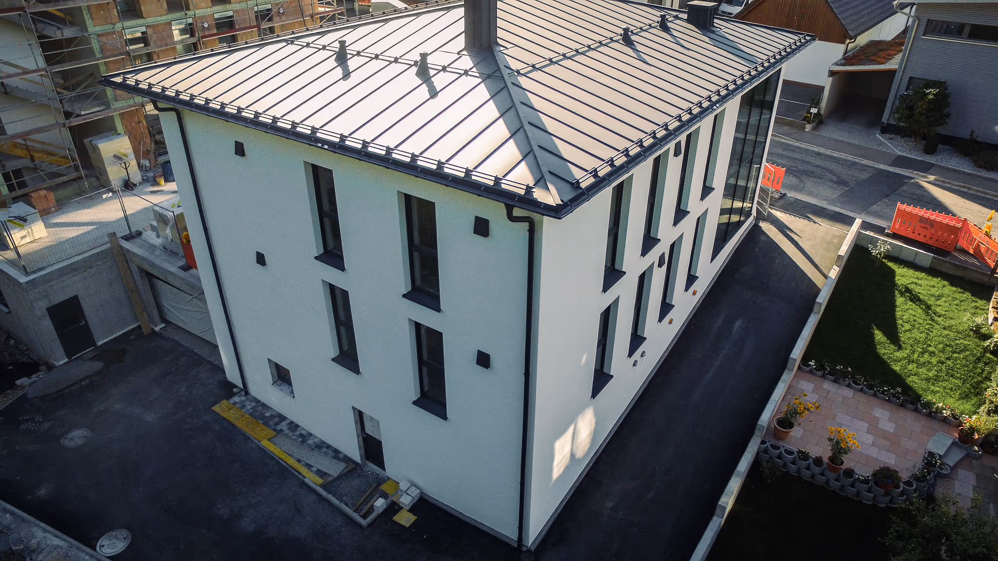 Modernes, weißes Mehrfamilienhaus mit dunklem Metalldach, frisch asphaltierter Zufahrt und angrenzendem Neubau mit Baugerüst.