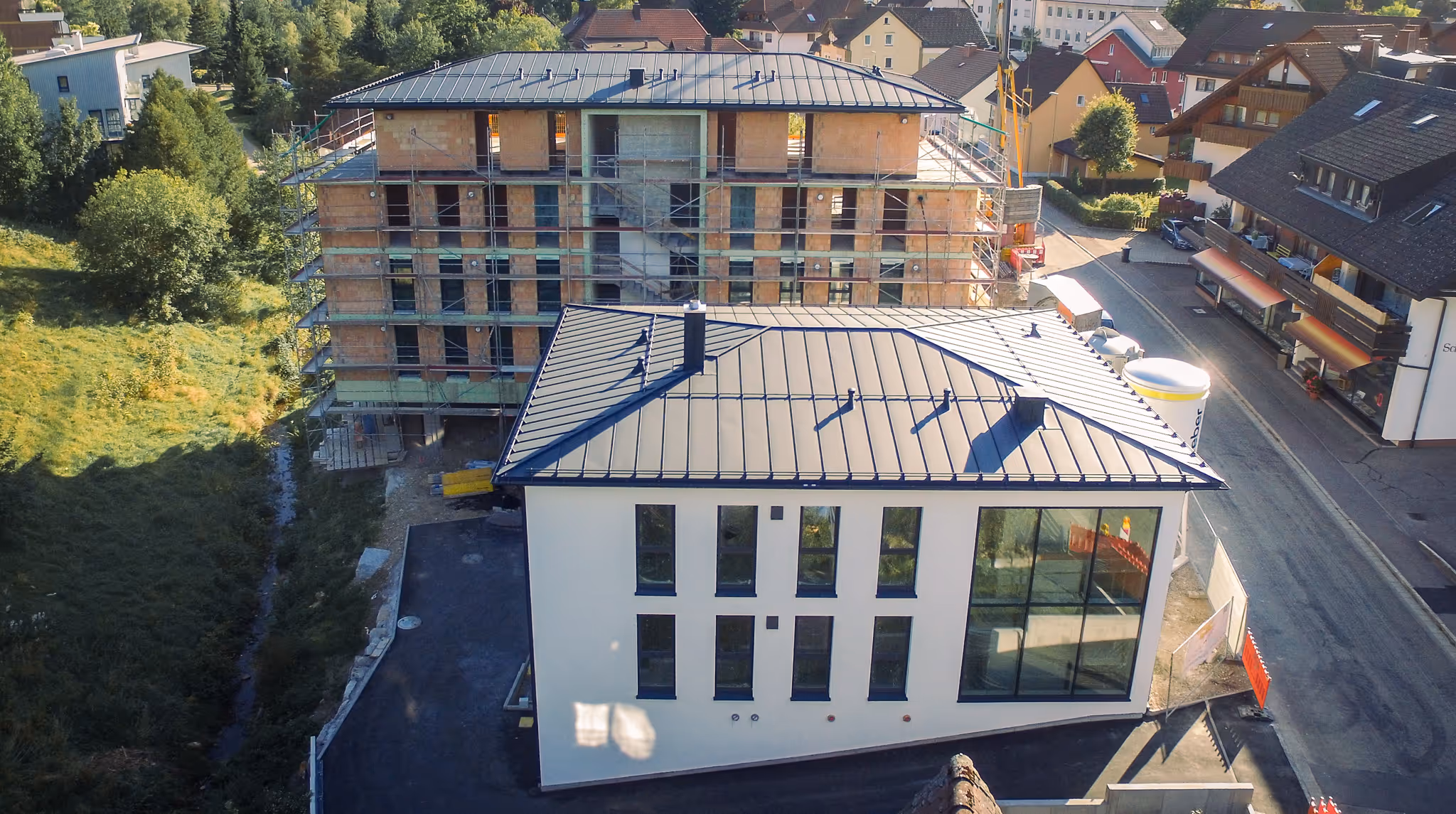 Neubau mit Glasfassade im Vordergrund, dahinter ein weiteres Gebäude im Rohbau mit Baugerüst, inmitten eines Wohngebiets.