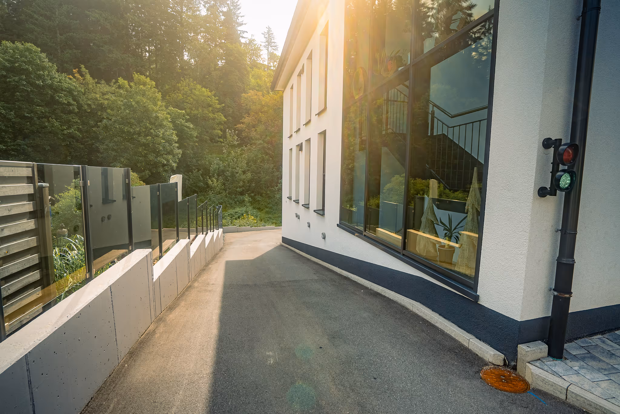 Moderne weiße Gebäudefassade mit bodentiefen Glasfenstern, umgeben von grünem Wald im Sonnenlicht.