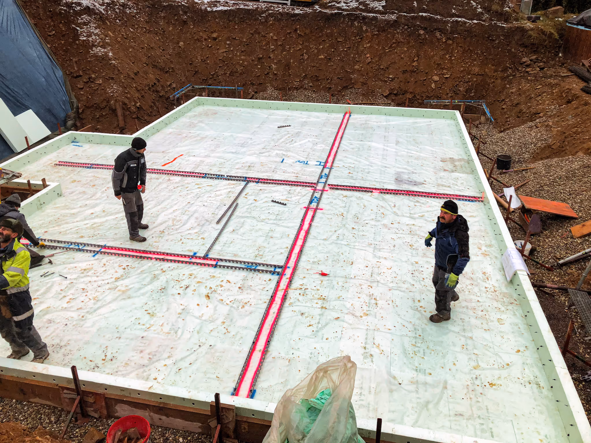 Zwei Arbeiter auf einem vorbereiteten Fundament mit Folienabdichtung und sichtbaren Leitungen.