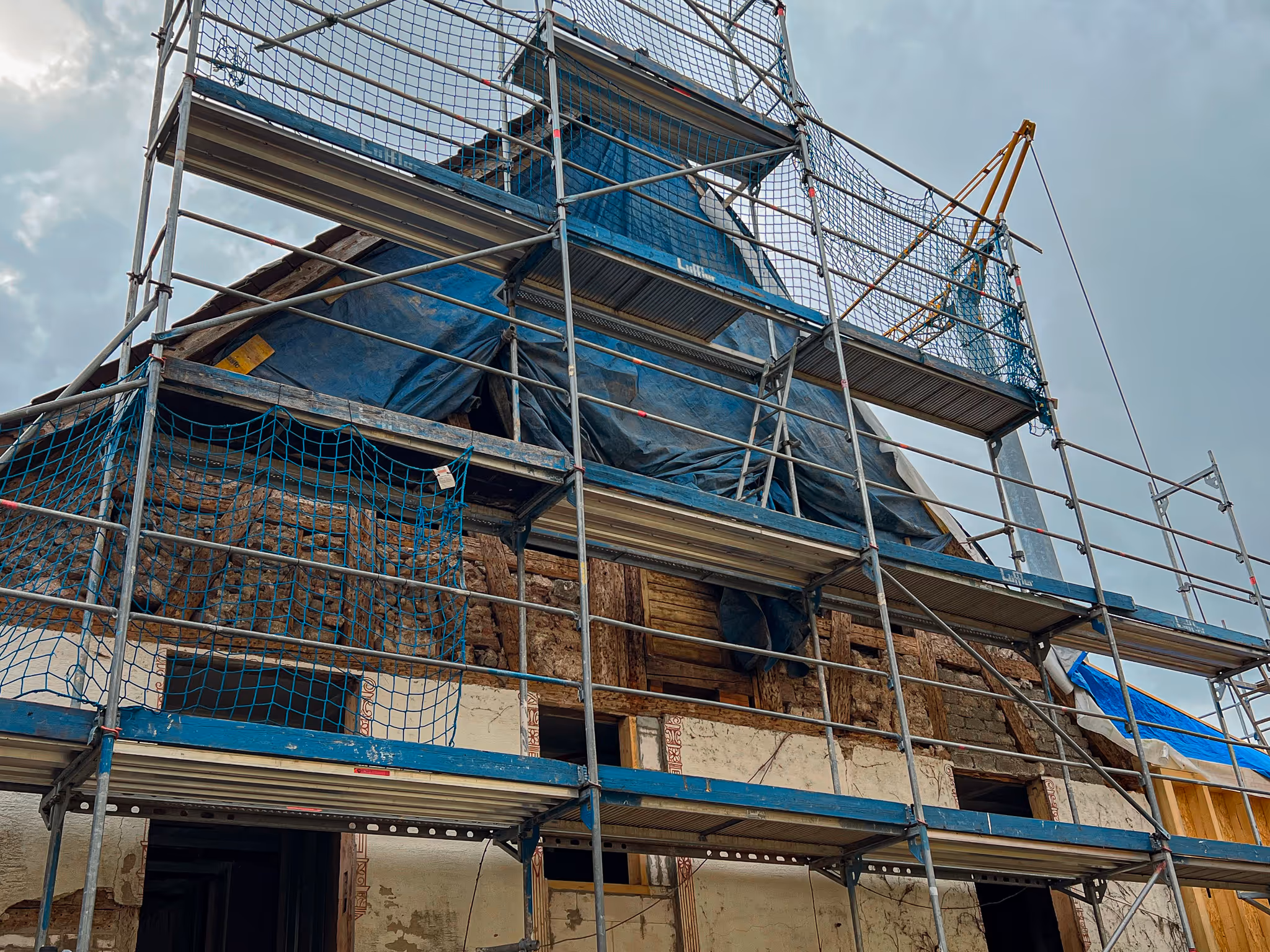 Außenansicht eines Gebäudes mit Baugerüst. Dach teilweise geöffnet und mit Planen abgedeckt, Fassade zeigt historische Lehmbauweise.