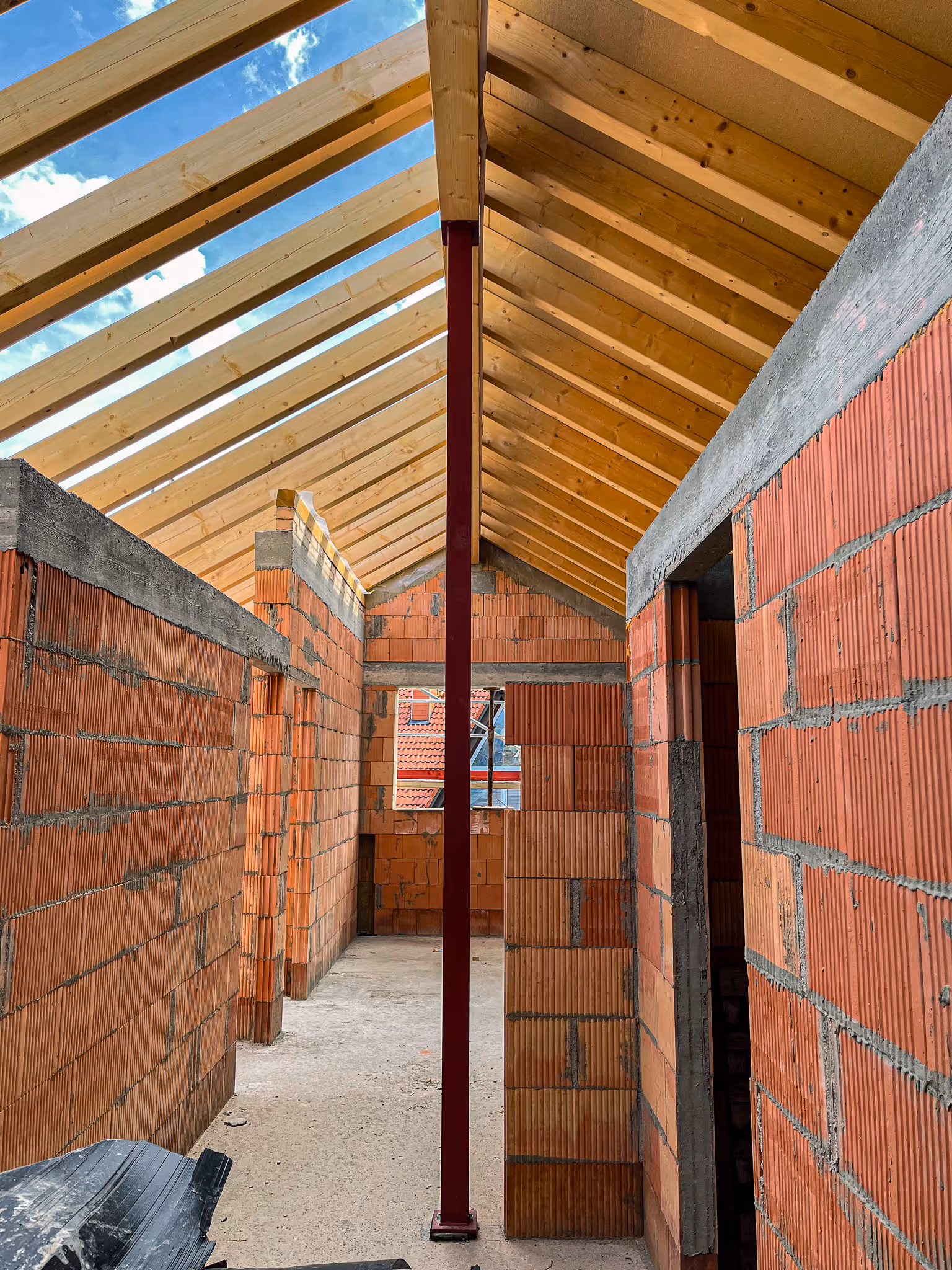 Blick in einen Rohbauflur mit gemauerten Ziegelwänden und Holzdachkonstruktion, durchzogen von einer mittig stehenden roten Stahlstütze.