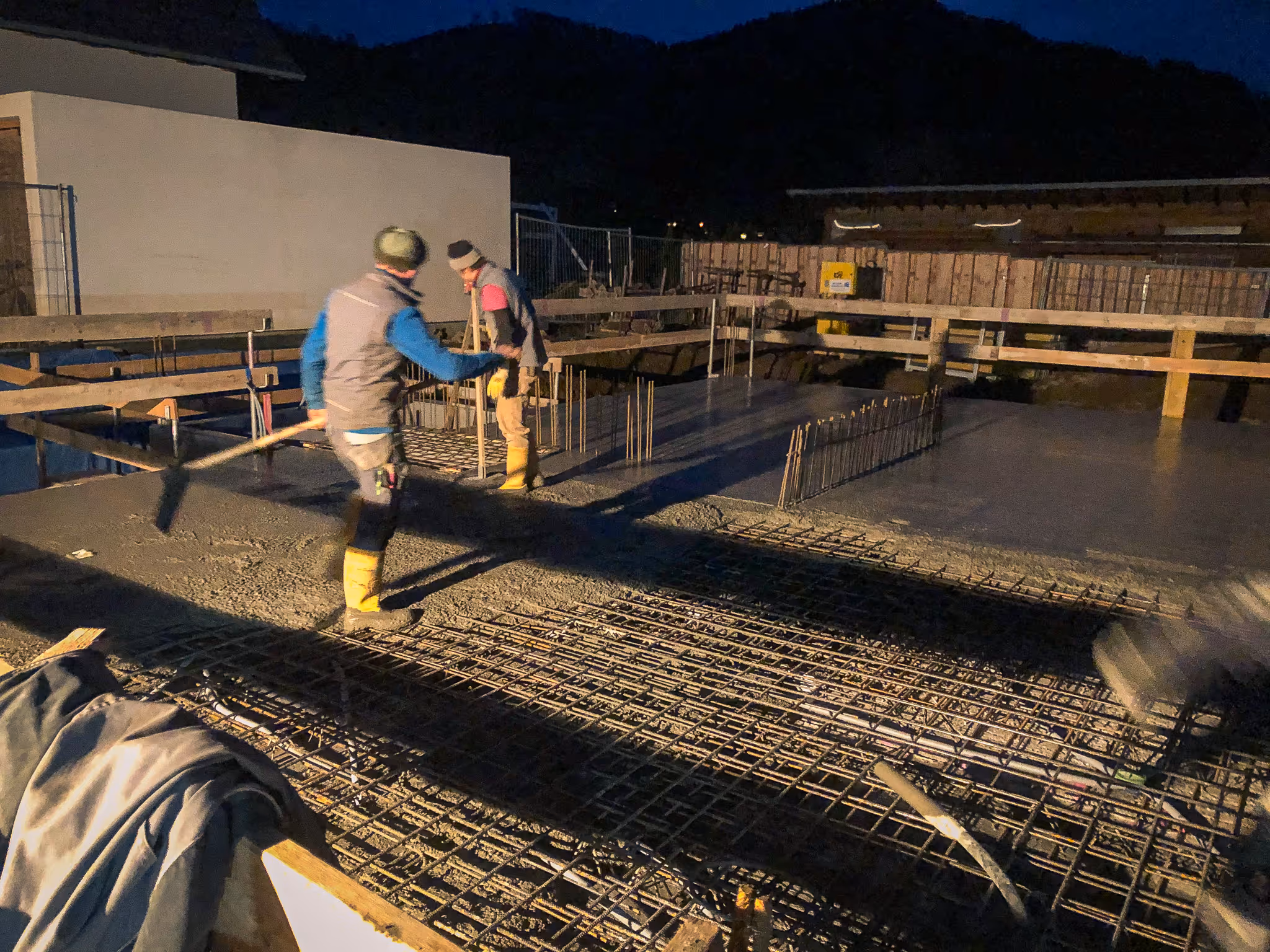 Zwei Bauarbeiter gießen und glätten Beton auf einer Baustelle bei Dunkelheit.