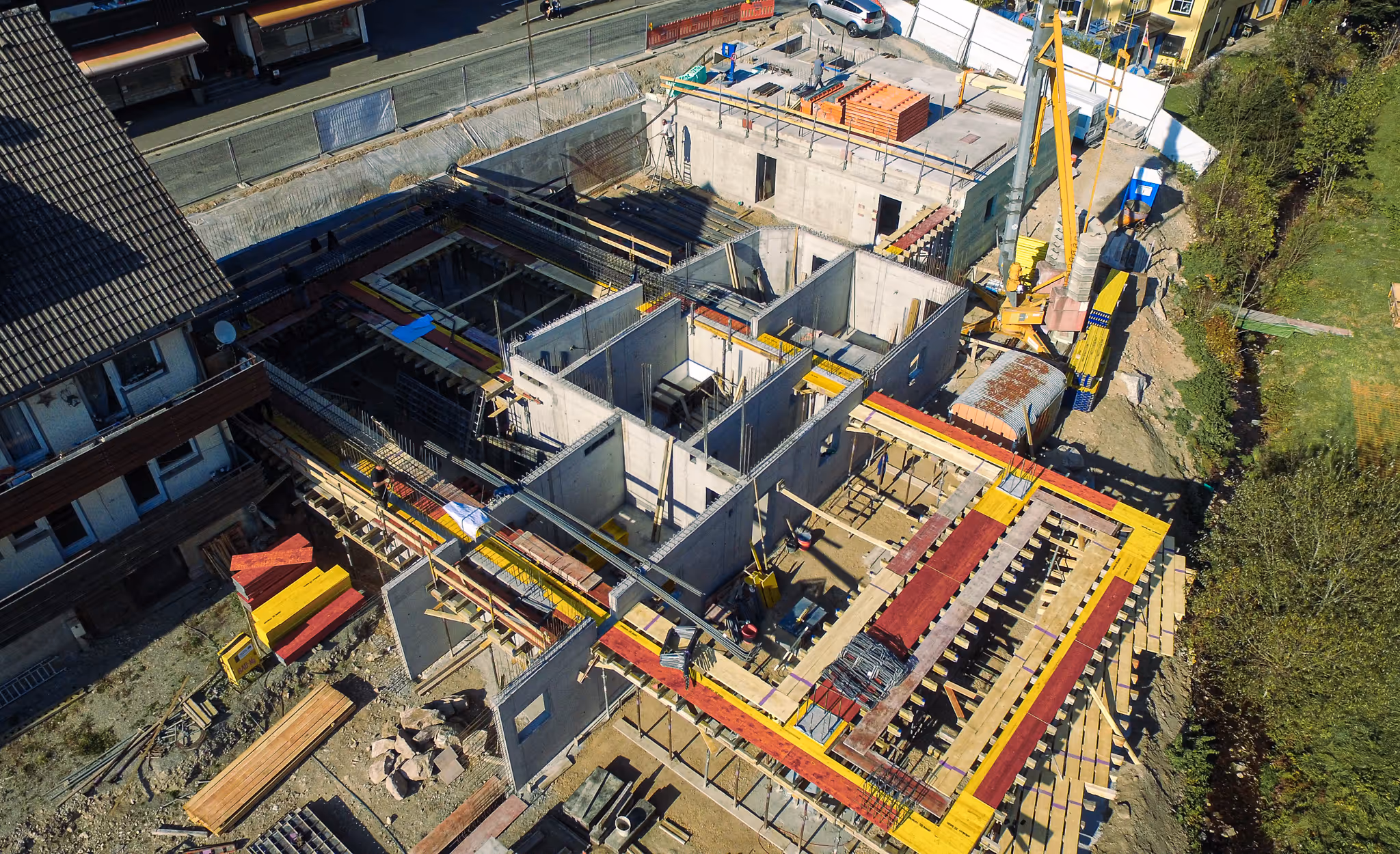 Luftaufnahme einer großen Baustelle mit Kran, Schalungen und ersten Betonarbeiten. Links angrenzend ein bestehendes Wohnhaus.