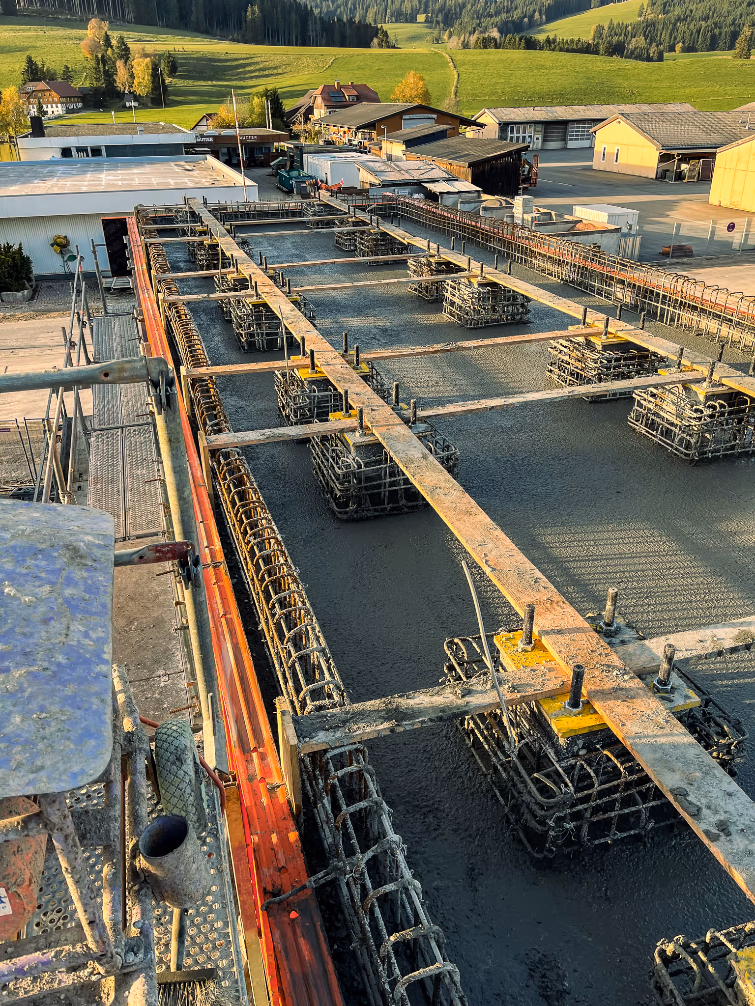 Luftaufnahme einer Baustelle mit frischem Beton und stahlbewehrten Fundamenten; im Hintergrund grüne Hügel und Gebäude.