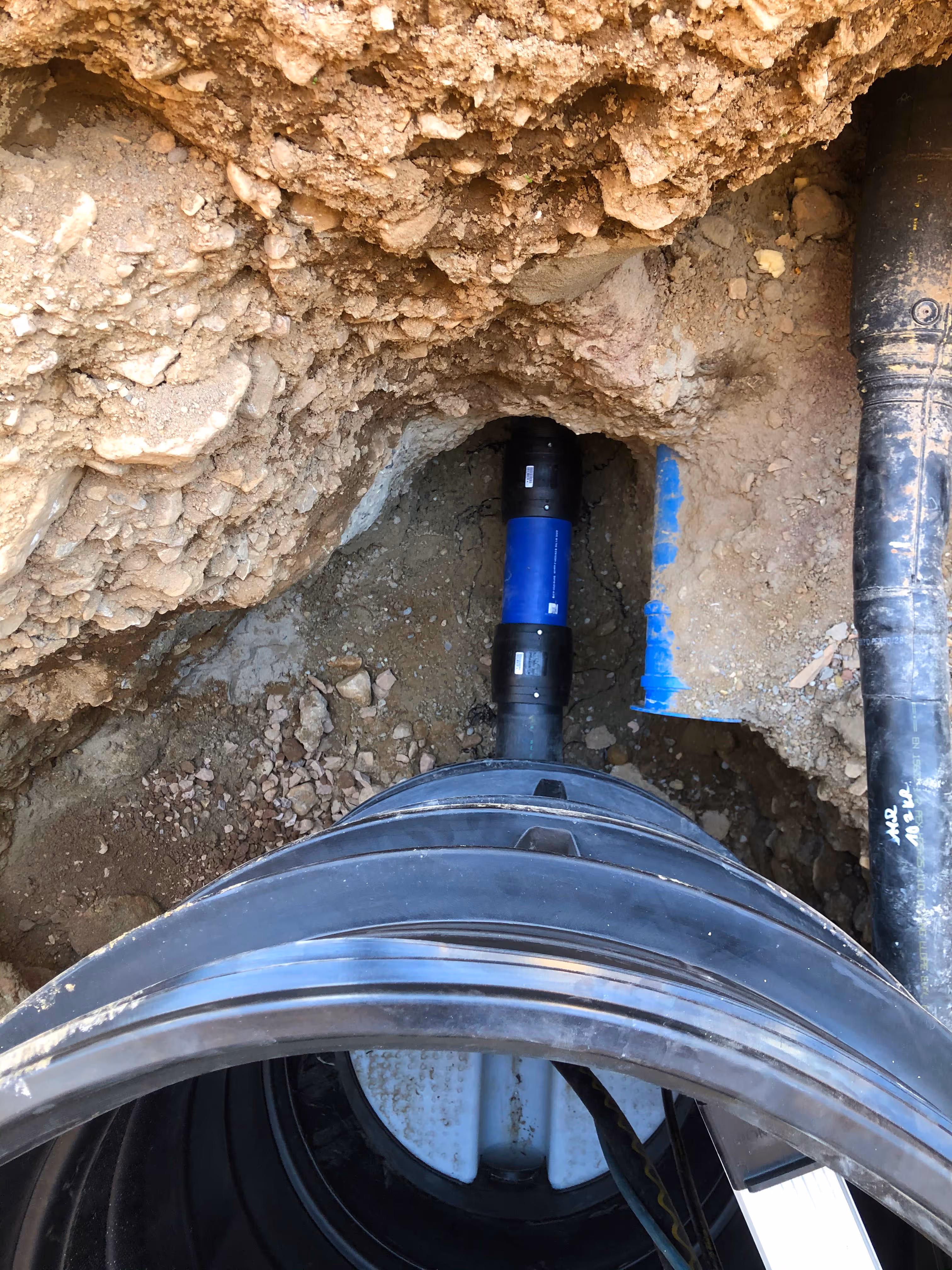 Blaue Wasserleitung in Baugrube zwischen Felsen und Erdreich, teilweise freigelegt.