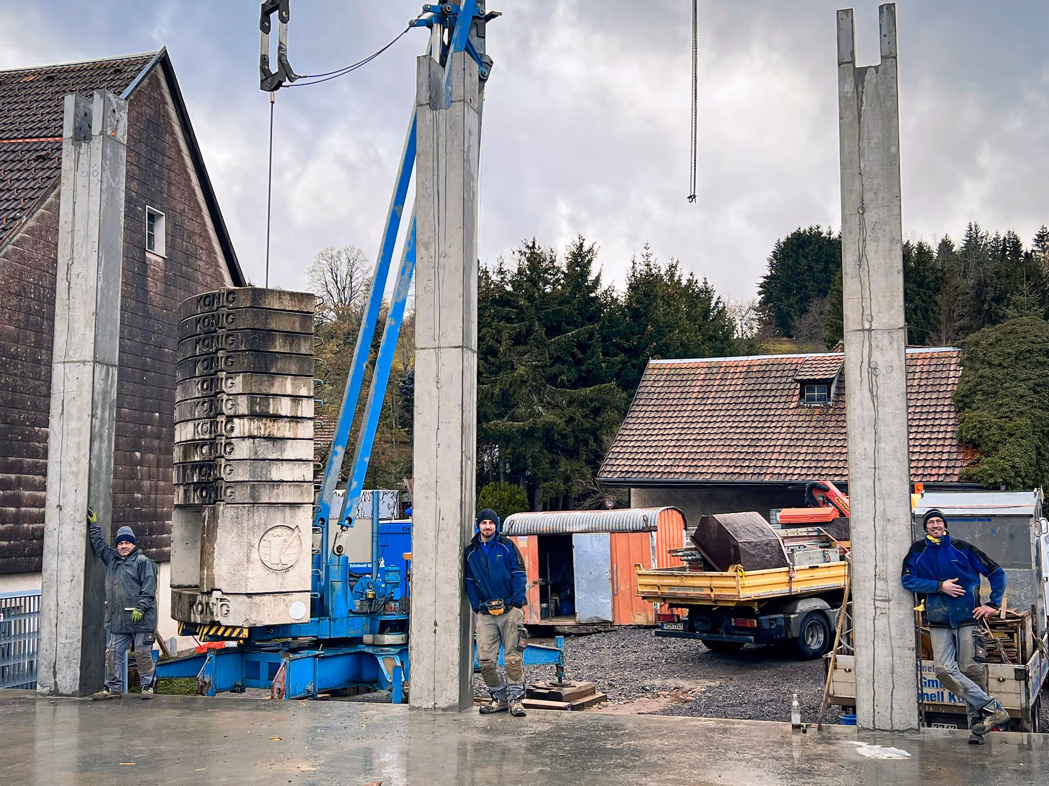 Drei Bauarbeiter stehen zwischen aufgerichteten Betonstützen auf einer Baustelle.